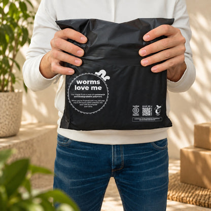 A person in a white sweatshirt and blue jeans holds a black compostable mailer labeled 'worms love me' in a bright, sunlit outdoor setting with plants and cardboard boxes in the background.