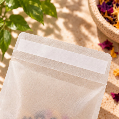 A translucent glassine bag with a blank white label strip, photographed on a sunlit beige surface with green leaves and dried flowers in the background.


