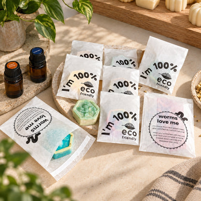 Several small white eco-friendly product pouches labeled 'I'm 100% eco friendly' and 'worms love me' are arranged on a beige stone surface with plants, amber bottles, and a green hexagonal bar of soap visible through one pouch.