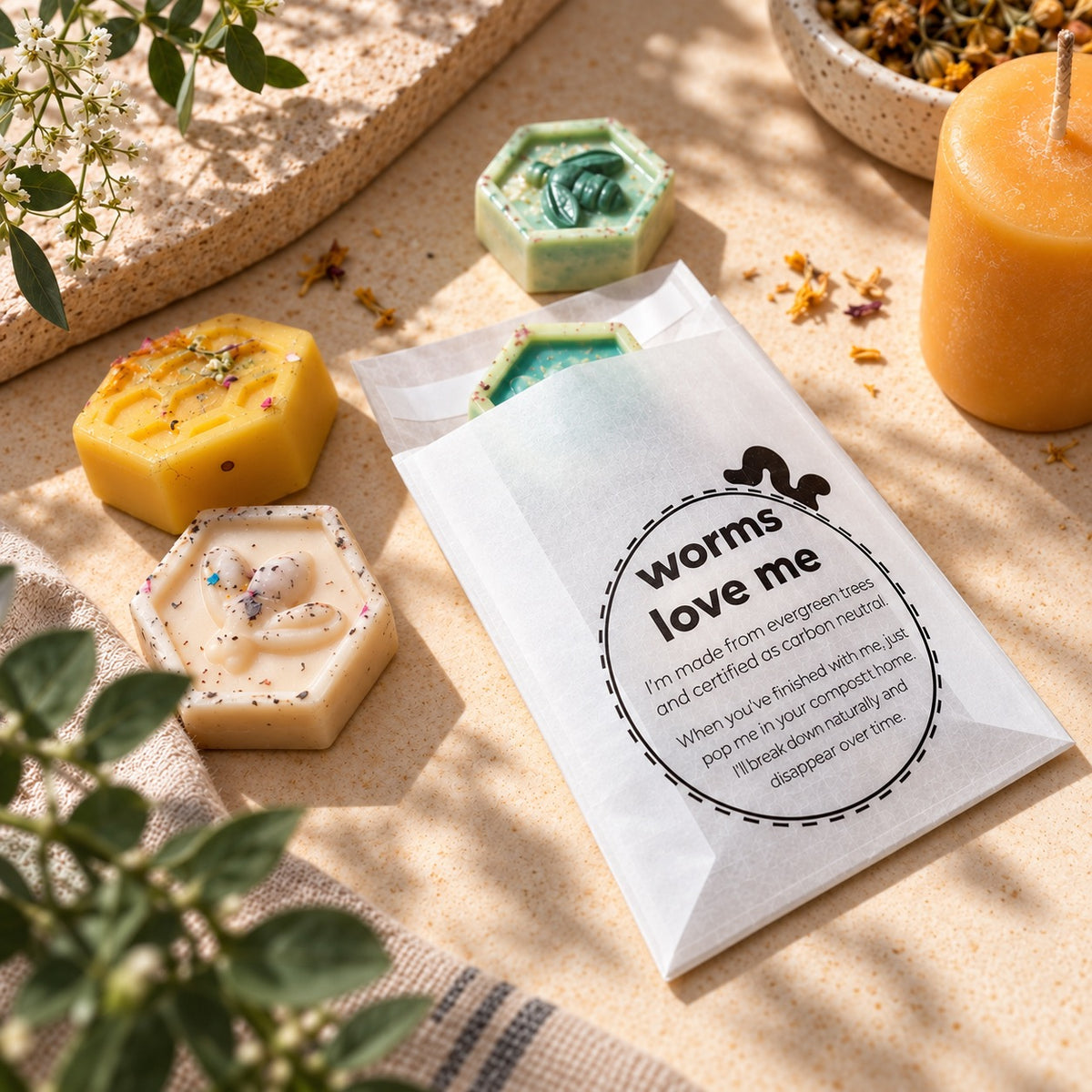 A white glassine paper bag labeled 'worms love me' with a small black worm illustration, placed on a beige textured surface with several hexagonal handmade soaps around it. The scene includes greenery, dried flowers, and a yellow candle in a real styled setting.