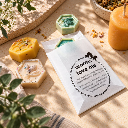 A white glassine paper bag labeled 'worms love me' with a small black worm illustration, placed on a beige textured surface with several hexagonal handmade soaps around it. The scene includes greenery, dried flowers, and a yellow candle in a real styled setting.