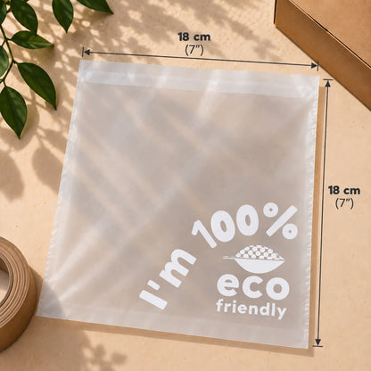 A translucent compostable poly bag with printed white text reading 'I'm 100% eco friendly' and dimension arrows showing 18 cm by 18 cm, styled on a beige surface with leaf shadows, tape, and a box corner visible.
