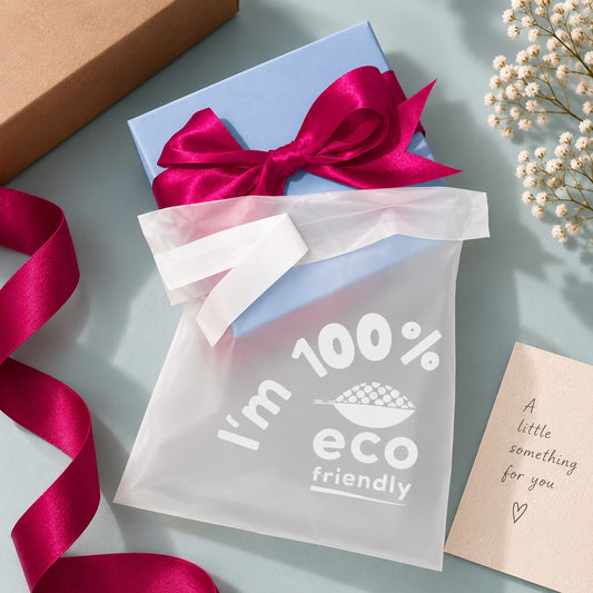 A gift box in a frosted translucent compostable poly bag printed with 'I'm 100% eco friendly'. The scene is styled on a pale blue surface with a brown kraft box corner, a pink ribbon loop, white baby's breath flowers, and a beige card reading 'A little something for you'.