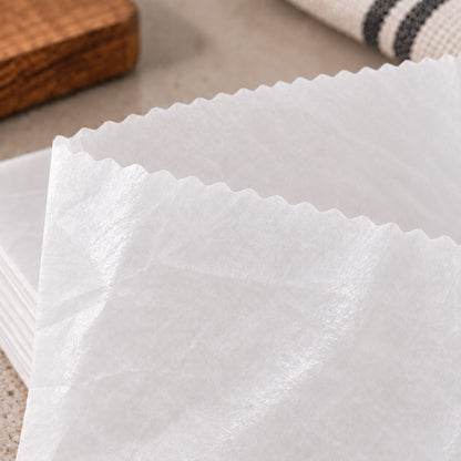 White scotchban greaseproof parchment paper sheets with scalloped edges stacked on a neutral surface, with a wooden block and a striped cloth partially visible in the background.

