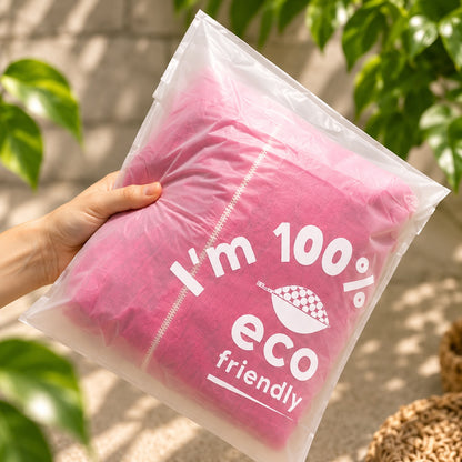 A hand holds a translucent compostable poly bag containing a bright pink pillow. The bag has white printed text reading 'I'm 100% eco friendly' with a small checkered leaf logo.