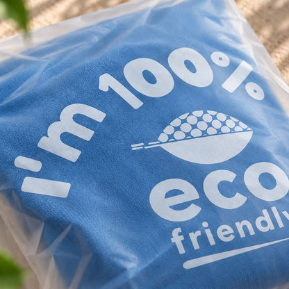 A folded blue towel or fabric item sealed in a compostable poly bag, printed with large white text reading 'I'm 100% eco friendly' and a white graphic of a bowl with dots.