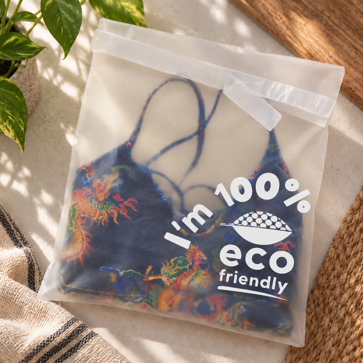 A semi-transparent compostable plastic bag containing a dark blue bikini top with colorful dragon prints, placed on a light surface with a plant and woven mat nearby. The bag has a printed white graphic that reads 'I'm 100% eco friendly'.