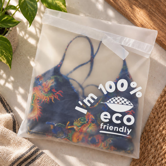 A semi-transparent compostable plastic bag containing a dark blue bikini top with colorful dragon prints, placed on a light surface with a plant and woven mat nearby. The bag has a printed white graphic that reads 'I'm 100% eco friendly'.