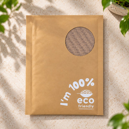 A brown kraft padded paper mailer envelope with a circular cutout window showing a textured brown insert, printed with 'I'm 100% eco friendly' in white text on a light stone surface with green leaves in the corners.