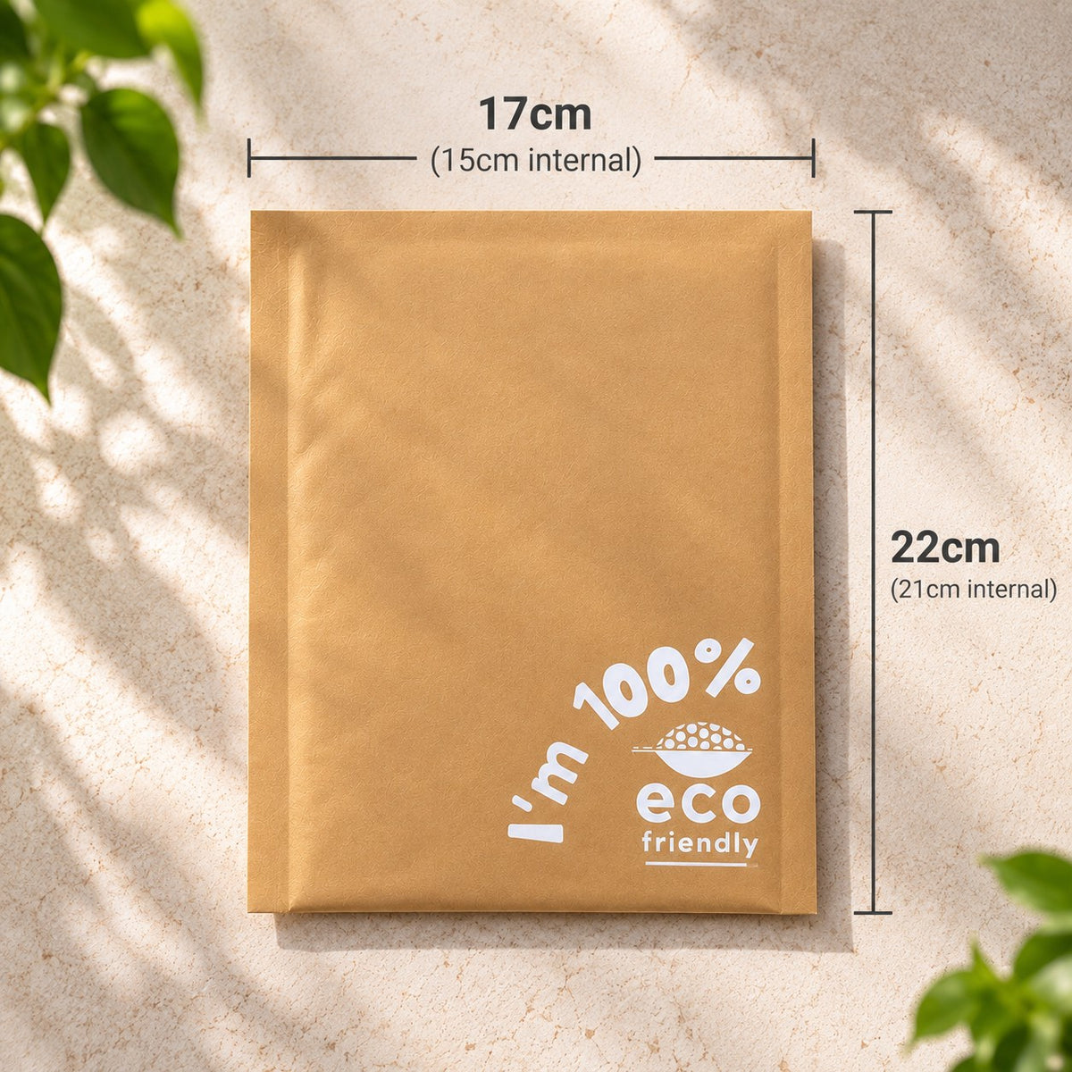 A brown kraft padded paper mailer envelope with white printed text reading 'I'm 100% eco friendly' and dimension callouts showing 17cm width and 22cm height, photographed on a light textured surface with leaf shadows.