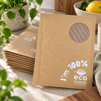 A stack of brown kraft paper pouches with a circular cutout window and white printed text reading 'I'm 100% eco friendly' and 'warm love me' on a countertop, styled with plants, a lemon, and a mug.