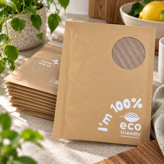 A stack of brown kraft paper pouches with a circular cutout window and white printed text reading 'I'm 100% eco friendly' and 'warm love me' on a countertop, styled with plants, a lemon, and a mug.