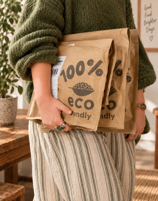A person in a chunky green sweater holds several brown paper mailing bags printed with '100% eco friendly' in a warm indoor setting.