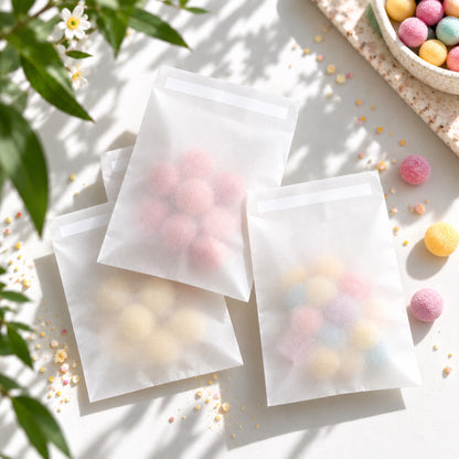 Three translucent white resealable glassine envelopes filled with pastel-coloured round candies, arranged on a bright white surface with soft shadows, decorative leaves, and scattered candy pieces.