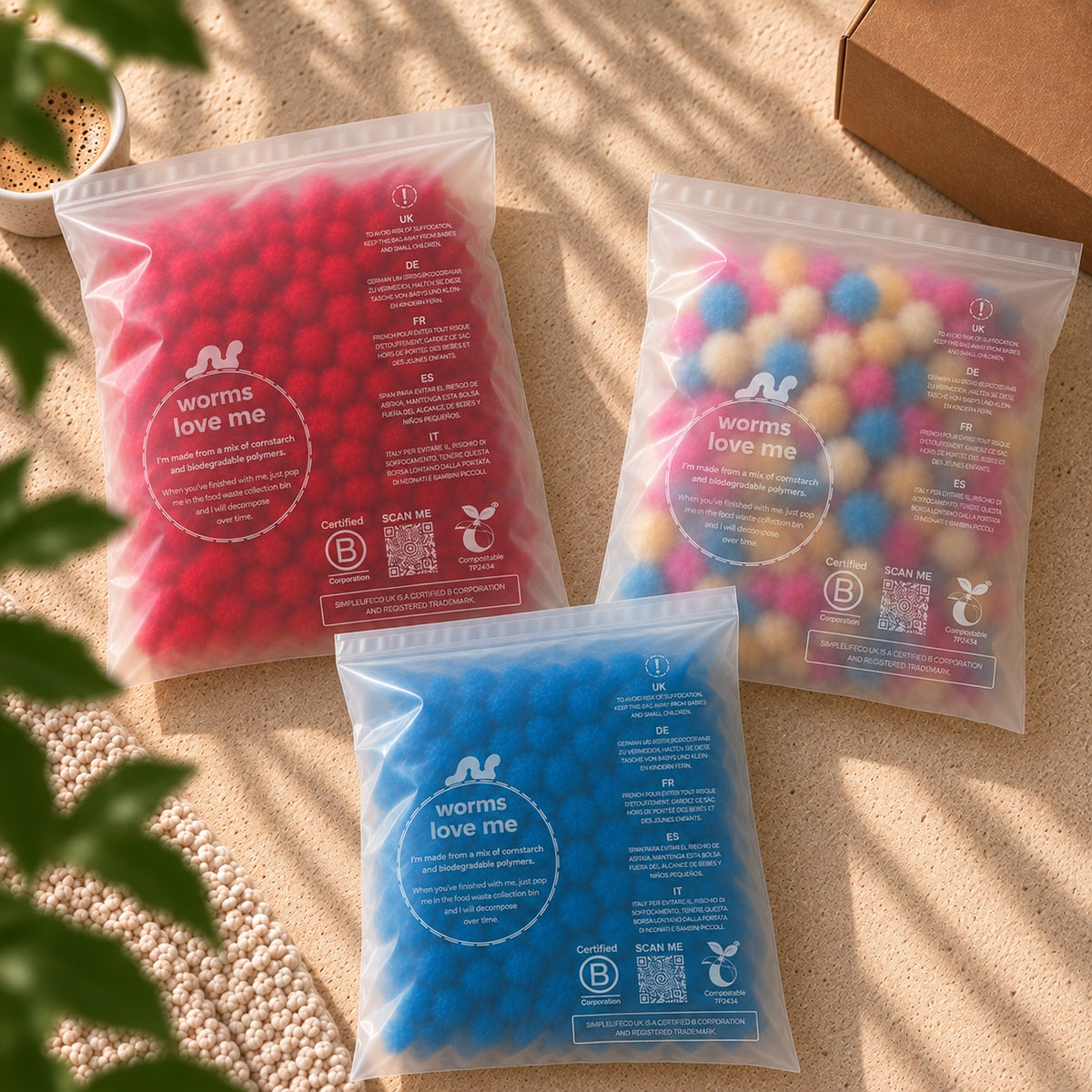 Three clear compostable poly bags filled with pom-poms are arranged on a textured beige surface with natural leaf shadows. One bag contains red pom-poms, one contains multicolour pom-poms, and one contains blue pom-poms.
