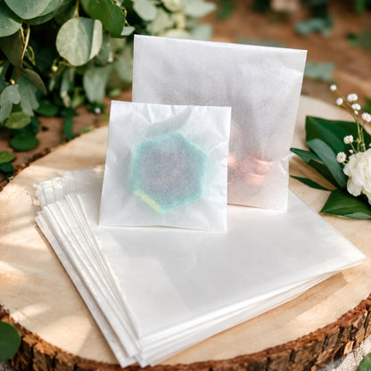 White paper glassine bags and a stack of white tissue-like sheets arranged on a round wood slice, with green leaves and small white flowers around them. One front bag is slightly translucent and shows a pale aqua-green hexagonal shape inside.