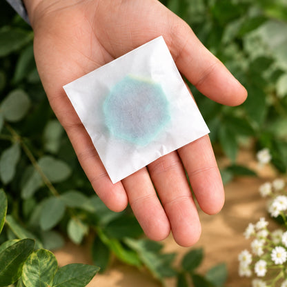 A hand holding a small white square glassine bag with a translucent aqua-blue circular centre, photographed against a blurred background of green leaves and small white flowers.