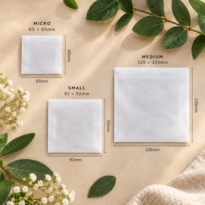 Three square white glassine paper bags labeled MICRO, SMALL, and MEDIUM with dimensions shown in millimetres, arranged on a beige textured surface with green leaves and small white flowers as styling props.