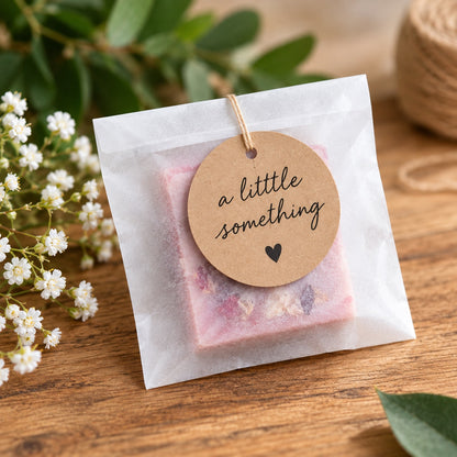 A small square bath soak or soap package in a translucent white glassine bag with a round kraft paper tag that reads 'a little something' and a small heart, placed on a wooden surface with flowers and greenery in the background.
