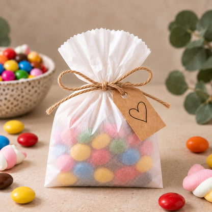 A small white drawstring candy bag filled with colourful round candies, tied with twine and a kraft paper tag with a heart, set on a beige surface with scattered candies and soft-focus background decor.