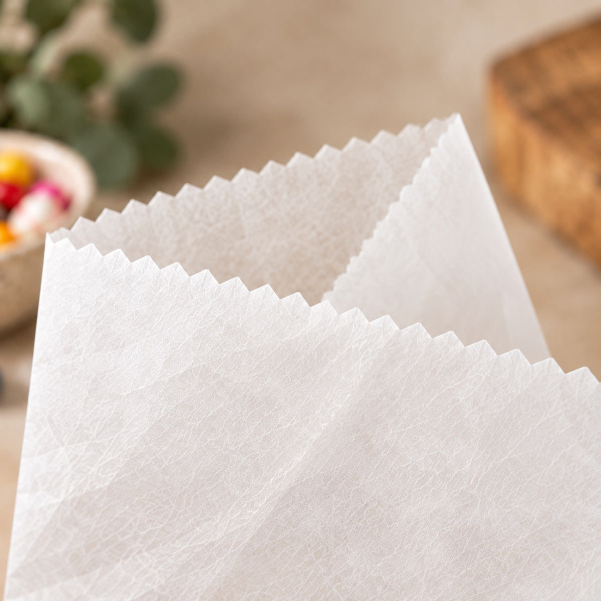 A close-up of a white textured greaseproof paper bag with crimped edges, shown against a softly blurred beige tabletop background with a small plant and wooden object in the distance.