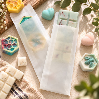 Two translucent white wax melts pouches glassine envelopes containing pastel-colored wax melts, styled on a beige fabric surface with decorative soap bars, hexagonal molds, and green leaves.